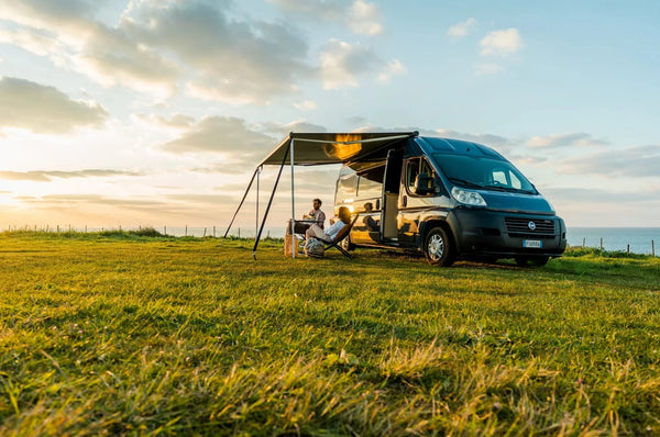 Fiamma wind-out awning extended on a campervan at sunset with people relaxing in camping chairs
