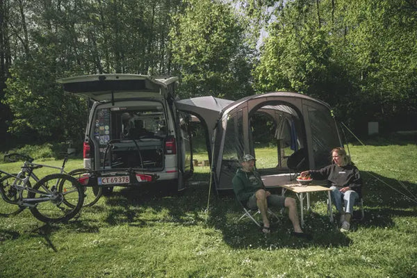 Outwell Armona drive-away awning attached to a campervan with two adults relaxing in camping chairs and bikes on a rear rack