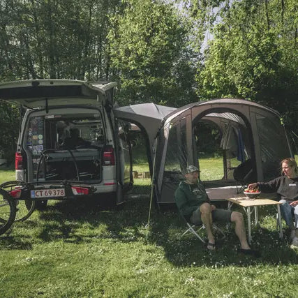 Outwell Armona drive-away awning attached to a campervan with two adults relaxing in camping chairs and bikes on a rear rack