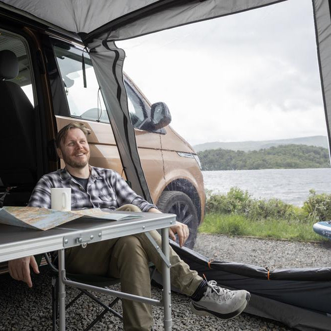 Man sitting in a drive-away awning attached to a campervan with chair, table and map