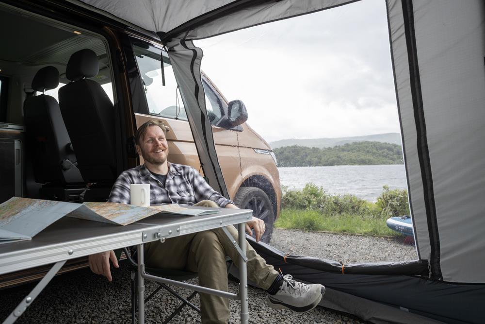 Man sitting in a drive-away awning attached to a campervan with chair, table and map