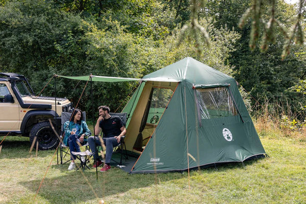 Coleman event shelter pitched beside an RV with people relaxing on camping chairs in sunshine and forest backdrop