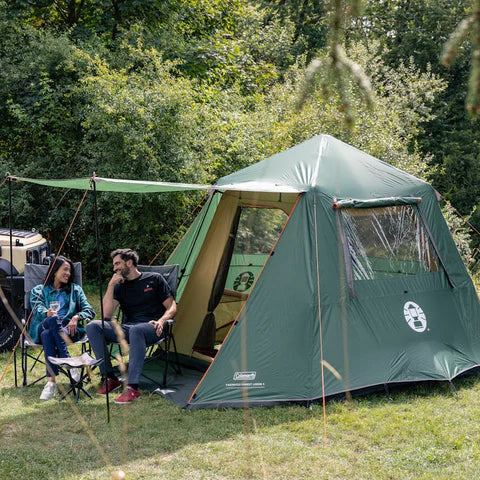 Coleman event shelter pitched beside an RV with people relaxing on camping chairs in sunshine and forest backdrop