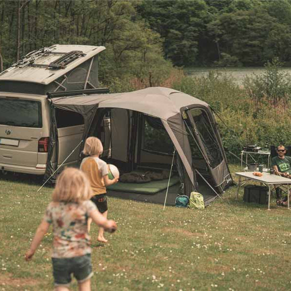 Outwell drive-away awning attached to a campervan with children running toward two adults seated in camping chairs at a table in sunshine