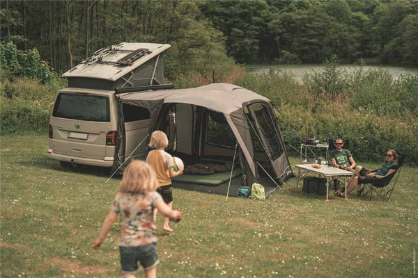 Outwell drive-away awning attached to a campervan with children running toward two adults seated in camping chairs at a table in sunshine