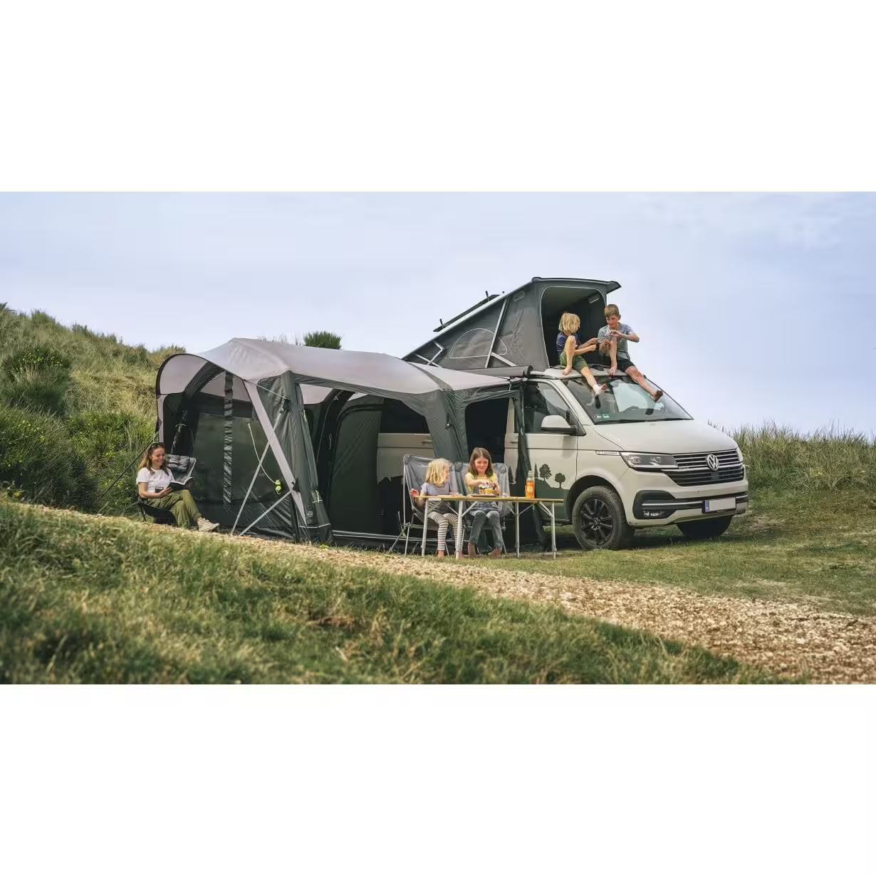 Outwell drive-away awning with full camping setup including campervan, family with four children and camping chairs in sunshine