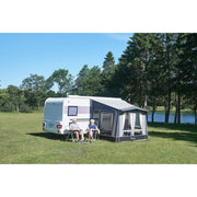 Two people sitting in front of a caravan with an awning on a grassy area near trees and water.