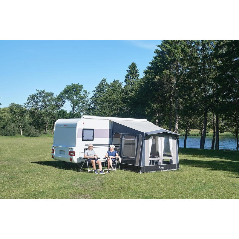 Two people sitting in front of a caravan with an awning on a grassy area near trees and water.